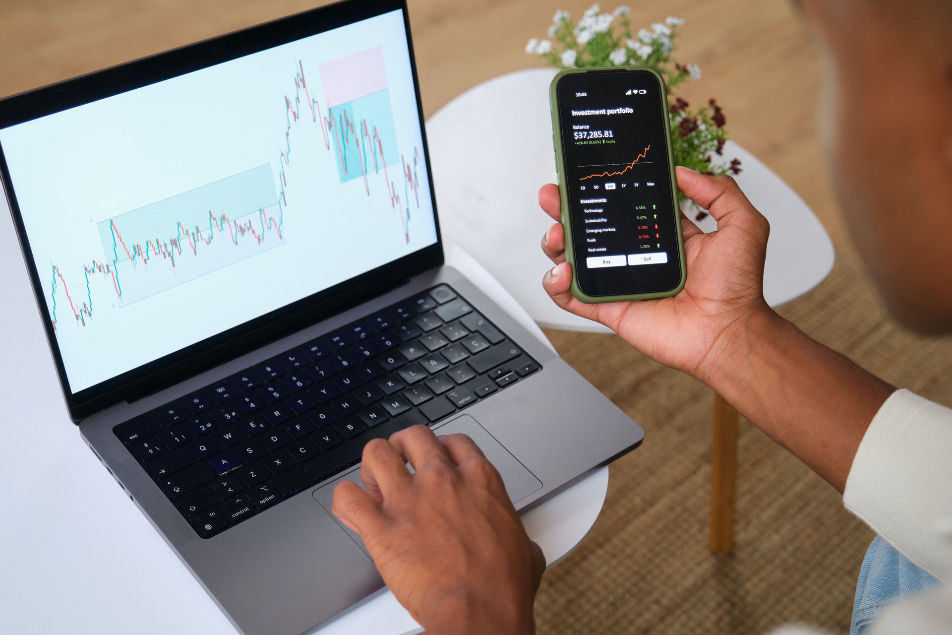 Trader analyzing charts on laptop and smartphone app at home office